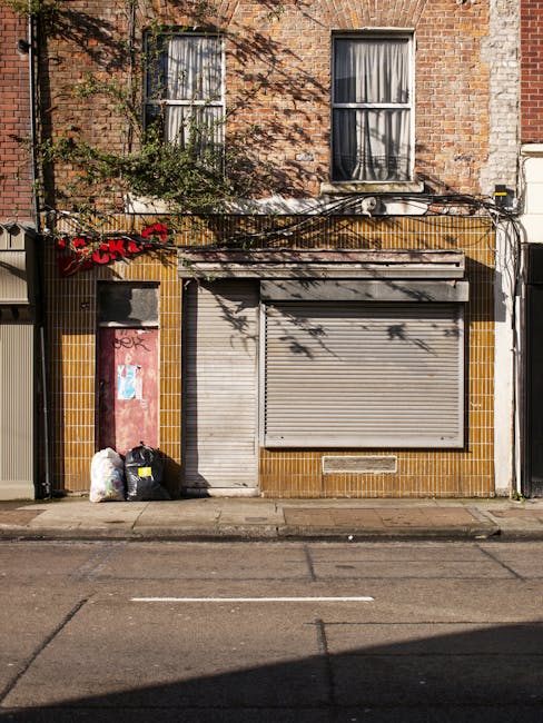 The image depicts the front of a small commercial building with a brick facade, featuring two vertically aligned windows with cream-colored curtains, positioned above the ground-level storefront. The lower section of the building is clad in brown and yellow tiles, with an aged, closed metal roller shutter covering the main entrance, and a separate narrow pedestrian door to its left. Near the base of the building on the pavement, there are three garbage bags, one white and two black, placed next to a small, boarded-up doorway. The pavement in front shows faint tire marks, and overhanging branches cast shadows onto the upper part of the facade. Exterior wires run horizontally across the building’s front, indicating typical urban infrastructure. The scene suggests an area where private or independent waste collection might take place, as evidenced by the visible rubbish bags outside, aligning with services provided by waste removal specialists such as Waste Removal Barnes.