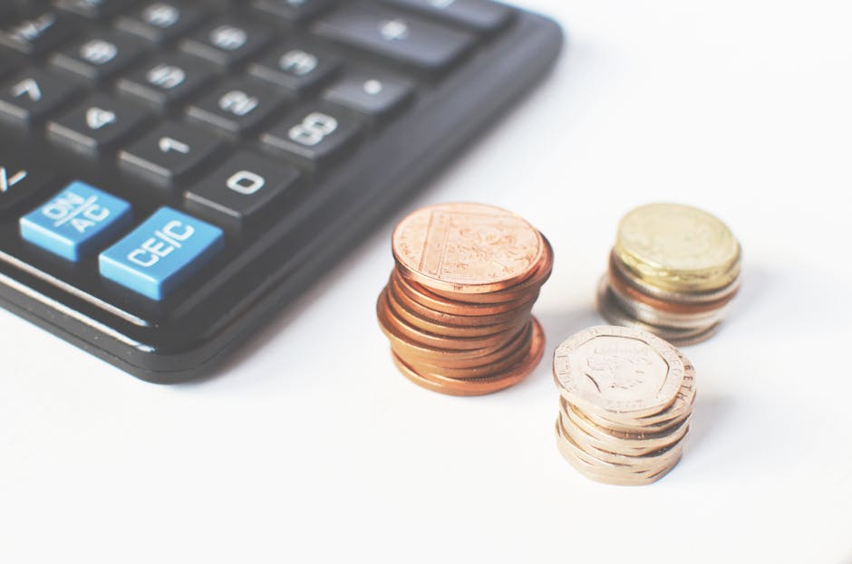 The image shows a close-up view of a black calculator with three stacks of British coins placed on a white surface. The calculator appears to be a basic model with visible numeric buttons, including a light blue 'AC', 'CE', and 'C' key on the left side. The coins include one copper penny stack, a smaller stack of silver coins, and a slightly larger stack of mixed coins, all arranged in neat piles. The copper penny coins have a reddish-brown finish with visible ridges along the edges, while the silver coins are lighter in color with some showing the familiar ridged edges of circulating currency. The background is plain white, emphasizing the coins and calculator. This scene may relate to the concept of budgeting, cost calculations, or financial planning, aligning with themes of costs associated with waste management services like rubbish removal provided by Waste Removal Barnes in the context of private waste handling or independent collection costs.