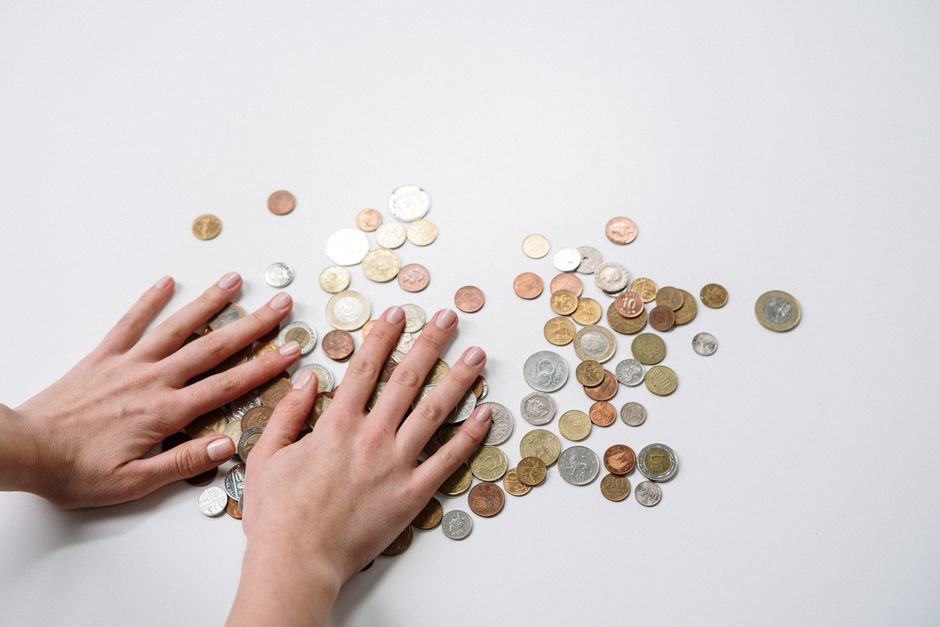 A pair of human hands with neatly trimmed nails and light skin tone are positioned in the lower left corner of the image, arranging or counting various coins scattered on a plain white surface. The coins are diverse in size, shape, and color, including silver, copper, and gold tones, indicating different denominations and currencies. The background is free of distractions, emphasizing the coins and the hands' subtle actions. The overall environment appears clean and well-lit, suggesting a context of financial sorting or counting, with no additional objects or environment features visible. This natural scene can be associated with financial management, cash handling, or the context of individual or private money collection, aligning with themes of independent disposal or supplementary cash transactions related to rubbish removal services, such as those offered by Waste Removal Barnes for uncomplicated, direct payment options.