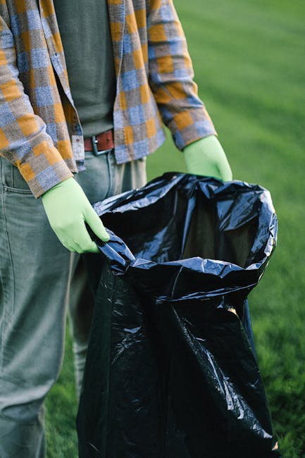 A person standing outdoors on a grassy area, holding open a large black rubbish bag lined with silver internal material. The individual wears a plaid shirt with shades of yellow, blue, and gray, over a dark green T-shirt, and has light green gloves on both hands. The background features a well-maintained lawn with blurred greenery, suggesting a garden or park setting. The person appears to be preparing or collecting garden waste for disposal, reflecting services related to private waste handling, rubbish removal, or on-site clearance provided by Waste Removal Barnes. The scene emphasizes the practical aspects of waste collection, with attention to detail in the material and textures of the bag, clothing, and gloves, typical of waste management activities aimed at environmentally responsible disposal outside of council-provided services.