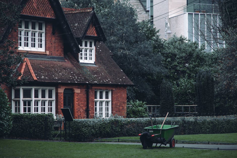 A green wheelbarrow with black handles and red wheels is positioned on a lawn in front of a red-brick house with a sloped, tiled roof. The house features white-framed windows, some divided into small panes, and a black front door. The wheelbarrow contains a wooden-handled garden tool, possibly a rake or hoe. Surrounding the house are neatly trimmed boxes and hedges, with a small black fence visible in front of the windows. In the background, there are modern high-rise buildings with glass facades and concrete structures, partially obscured by trees and shrubs, indicating an urban residential area. The scene appears to be set during daytime with natural lighting, and the overall environment suggests a maintained garden or front yard in a mixed urban-suburban setting. The presence of the wheelbarrow and garden tool subtly emphasizes garden maintenance or outdoor waste clearance activities, aligning with the theme of rubbish removal services such as those offered by Waste Removal Barnes.