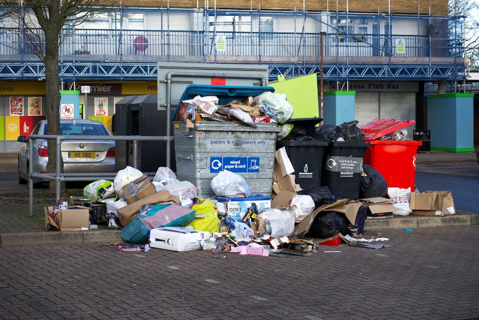 A large collection of overflowing rubbish bins and scattered waste bags situated on a paved sidewalk in front of a commercial area. The collection includes a grey recycling bin labeled for mixed paper and cardboard, a black general waste bin, and a bright red bin, all positioned adjacent to each other with their lids open. Surrounding the bins are numerous cardboard boxes, plastic bags, and loose paper, some of which are crumpled or torn. The waste spills onto the ground, mixing packaging, paper, and assorted refuse, indicating an unscheduled or excess collection. Behind the waste, there is a street scene with parked cars, including a silver vehicle, and a multi-storey building with a blue metal scaffolding structure and green trim around the storefronts. The environment appears urban, with a tree visible on the left side, and the lighting suggests overcast weather conditions. This scene highlights the importance of waste management services such as those offered by Waste Removal Barnes for efficient, alternative rubbish removal outside local authority bin collection cycles, especially in busy commercial or residential areas.
